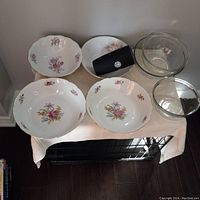 Top-down view of 4 porcelain bowls with floral patterns, 3 clear glass bowls, and 1 black thermos arranged on beige cloth surface