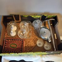 Top-down photo of the kitchenware lot inside a box showing the glass trays, ceramic shakers, metal gravy bowl, glass sugar bowl, metal paper towel holder, straw mats and glass cups.