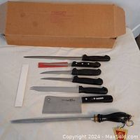 Photo shows 6 knives and one steel rod sharpener laid out next to a cardboard box on a white table surface. Knives have black or red handles and include various styles such as carving knives, paring knife, and cleaver.