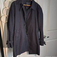 Full frontal image of the black men's raincoat hanging on a door showing length, collar, and button closure.