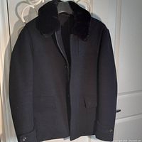 Full view of the men's black wool jacket with black fur collar hanging on a white hanger. Shows front and top of jacket.