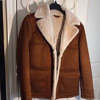 Front view of brown suede men's jacket with white fur lining on collar and inside hanging on a black hanger