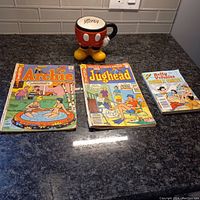 Overview of all items on dark countertop under kitchen cabinet light: three comic books and Mickey Mouse ceramic cup.