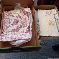 Two boxes each containing a folded tablecloth; left box holds 84 x 68 inch tablecloth with red French toile motif, right box holds 100 x 68 inch cream-colored embroidered tablecloth and a set of 12 napkins.
