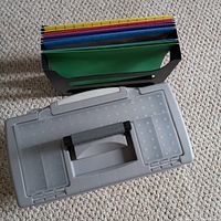 Overview of black hanging file organizer containing multiple colored folders and closed light grey plastic document holder on carpeted floor.