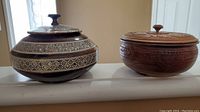 Photo showing two carved wooden bowls with lids on a surface, one with white inlay and the other with carved floral wood pattern.