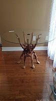 Side view showing clear round glass top resting on a detailed camel-shaped wooden base on hardwood floor.