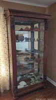 Front view of tall wood china/display cabinet with glass shelves and items inside. Carved wood accents on corners visible.