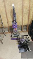 Full view of Dyson DC14 Animal upright vacuum, attachments in basket, and manual on floor, set against unfinished basement wall.