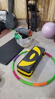 Wide view showing aerobic stepper, hula hoop, storage bin with yoga mats, and pink balance ball in basement setting.