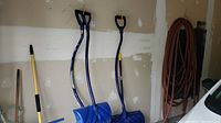 Three blue snow shovels with wide blades and D-handles hanging on garage wall with coiled orange-brown garden hose.