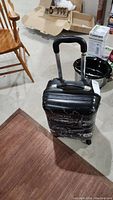Black Heys hardcover suitcase with white sketches, extendable handle, and wheels on cement floor near chairs and kitchen items.