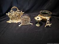 Photo showing five brass decorative items including napkin holders, incense holder, plate holder, and plant holder, all displayed against a dark background.