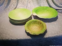 Photo of 8 green ceramic luncheon plates designed as lettuce leaves stacked, with small and large bowls shaped like lettuce leaves in matching green and yellow-green colors.