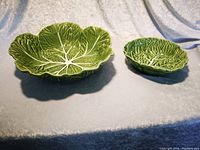 Photo showing two ceramic cabbage leaf salad bowls, one large and one small, placed on a light fabric background.