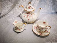 Photo showing porcelain teapot, creamer, and teacup with saucer on fabric backdrop, decorated with pink rose floral patterns and gold trim.
