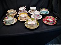 Photo showing 9 teacups and saucers with varying floral and gold detailed designs plus 1 extra cup and 1 extra saucer, all displayed on black cloth background.
