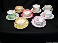 Eight assorted porcelain tea cups and matching saucers with various floral patterns and colored trims, displayed on black background