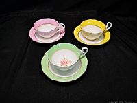 Three Bell China tea cups and matching saucers in yellow, green, and pink colors with floral and gold trim designs, arranged on a black background.