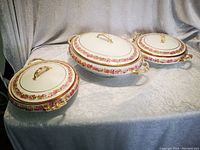 Three covered serving dishes displayed on grey velvet backdrop: two round dishes and one oval soup tureen.