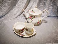 Photo showing Royal Albert Moss Rose teapot with lid alongside creamer and sugar bowl on an oval plate, all white porcelain with pink/red rose floral decoration and gold trim.