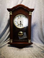 Front view of Hermle wooden wall clock with curved top wooden case, glass front revealing clock face with Roman numerals and brass pendulum below. Key and instruction papers are visible inside.