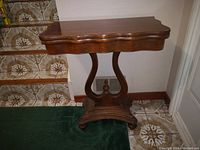 Wooden hall table with scalloped-edge swivel top, curved central support, and carved base shown near tiled stairs and wall.