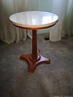 Photo of a small round wooden end table with a white marble top, turned pedestal base, and four feet standing on carpeted floor with curtains in background.