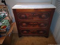 Front view of a medium-tone wooden small dresser with three drawers and decorative carved wood panels on each drawer. The top is covered with a white marble slab with subtle gray veining. The dresser has a scalloped front bottom edge and stands on short rounded feet.