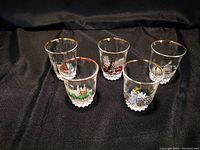 Photo showing set of five clear shot glasses arranged on black cloth, each with gold rim and different British landmark image.