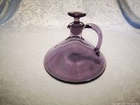 Wide bottom medium purple glass decanter with stopper lid and handle placed on a white surface with fabric background.