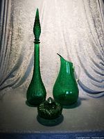 Photo showing the tall slender green glass decanter with pointed hobnail stopper, matching green glass pitcher with clear handle, and small green glass covered candy dish with textured pattern, all on a soft textured backdrop.