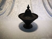 Black glass covered dish with textured surface and pointed finial lid, shown upright on a light background.