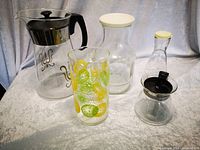 Five glass containers arranged on a light surface with grey curtain background. Includes coffee carafe, teapot, juice glass, pitcher, and dressing bottle.