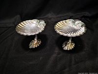 Two silver-tone footed shell-shaped soap dishes photographed on a black fabric background showing their ornate scallop shell foot supports and ridged shell plates.