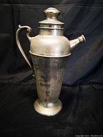 Silver plated vintage cocktail shaker with ornate handle, spout, and lid showing age wear and patina.