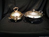 Photo showing two covered serving dishes with handles and feet on a black background, both exhibiting tarnishing.