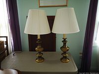 Two brass table lamps with cream pleated fabric shades on a plastic table in a room with green walls and curtains.