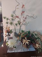 Four assorted faux flower pieces on a table including green pine-like stems, peach and white flowers, and mixed botanical elements in glass and ceramic containers.