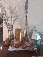Six decorative items on a table including metallic vases, a wooden vase, a glass lantern, and candle holders in amber glass with metal decoration.