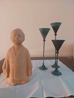 Photo showing ceramic monk figurine and three metal candle holders on table against wall.