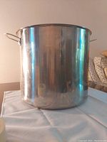 Image of a large stainless steel stock pot with two side handles on a white cloth surface, neutral background.