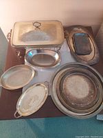 Photo of silver plated serving items including a rectangular chafing dish with lid, multiple large round platters, and various oval serving platters with handles on a wooden surface.