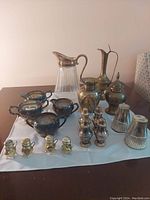 Photo showing assorted silver plated salt shakers, pitchers with glass and silver plated bodies, bowls, and other small table items arranged on a table with a white cloth.
