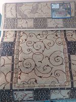 Photo of folded beige area rug with brown scroll patterns and dark blue borders, showing brand label attached.