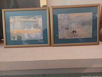 Photo showing two framed Renoir art prints side by side on a beige surface against a wall. Both frames have a green mat and light wood frame.