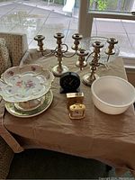 Overview of the nine pieces including silver-plated candelabras, clocks, milk glass bowl, and decorative bowls and plates on a table indoors by window.
