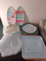 Photo showing eight assorted serving platters on a table, including floral ceramic platters, striped and floral patterned plastic platters, and clear plastic platters, some wrapped in plastic.