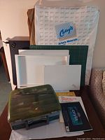 Photo showing wrapped canvas from Curry's Artist's Materials, multiple white canvases, a green cutting mat, green plastic storage box, and two paper pads on a table by a sofa and cabinet.