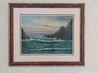 Framed Braunstan seascape painting showing ocean waves crashing between rocky cliffs under an overcast sky.